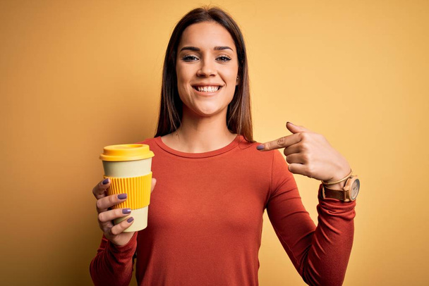 Jeune belle femme brune buvant une tasse de café à emporter sur fond jaune avec un visage surprise pointant du doigt vers lui-même
 - Photo, image
