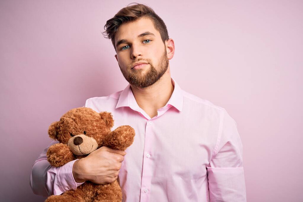 Fiatal szőke férfi szakáll és kék szemek gazdaság teddy maci játék felett rózsaszín háttér egy magabiztos kifejezés okos arc gondolkodás komoly - Fotó, kép