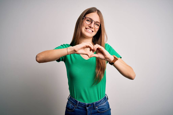 Young beautiful redhead woman wearing casual green t-shirt and glasses over white background smiling in love doing heart symbol shape with hands. Romantic concept. - Valokuva, kuva