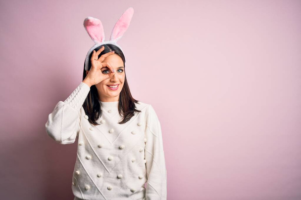 Joven mujer caucásica con lindas orejas de conejo de Pascua sobre fondo rosa aislado haciendo buen gesto con la mano sonriente, ojos mirando a través de los dedos con la cara feliz
. - Foto, Imagen