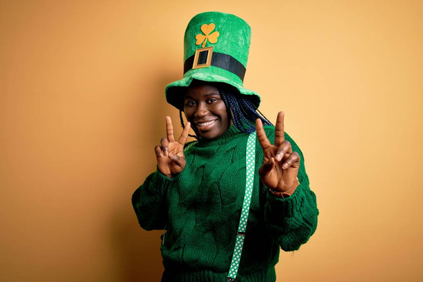 Плюс розмір афро-американської жінки з плечима в зеленому капелюсі з конюшиною на патриках день посміхається, дивлячись на камеру з пальцями, які роблять знак перемоги. Номер два. - Фото, зображення