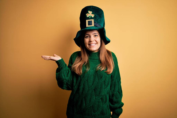 Jovem bela mulher morena usando chapéu verde em st patricks celebração do dia sorrindo alegre apresentando e apontando com palma da mão olhando para a câmera
. - Foto, Imagem