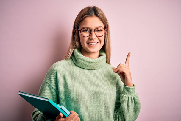 Young beautiful woman wearing glasses holding books over isolated pink background surprised with an idea or question pointing finger with happy face, number one - Foto, immagini