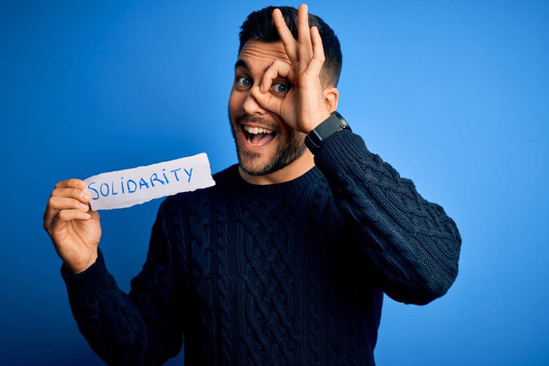 Young handsome man holding paper with solidarity message over blue background with happy face smiling doing ok sign with hand on eye looking through fingers - Photo, Image