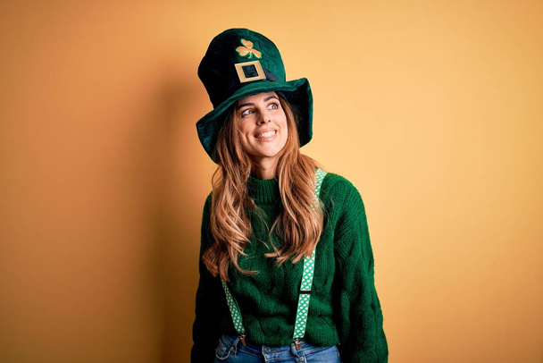 Mulher morena bonita usando chapéu verde com trevo celebrando dia santo patricks olhando para o lado com sorriso no rosto, expressão natural. Rindo confiante
. - Foto, Imagem