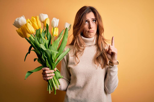 Young beautiful brunette woman holding bouquet of yellow tulips over isolated background Pointing up looking sad and upset, indicating direction with fingers, unhappy and depressed. - Photo, Image