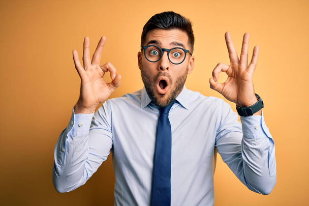 Young handsome businessman wearing tie and glasses standing over yellow background looking surprised and shocked doing ok approval symbol with fingers. Crazy expression - Photo, Image
