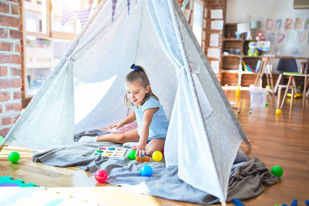 Giovane bella ragazza bionda ragazzo godendo di giocare a scuola con i giocattoli all'asilo, sorridente felice giocando all'interno tenda indiana a casa
 - Foto, immagini