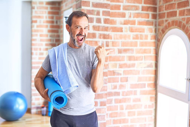 Красивый спортсмен средних лет с ковриком и полотенцем стоя перед делать упражнения в тренажерном зале указывая и показывая с большим пальцем в сторону со счастливым лицом улыбается
 - Фото, изображение