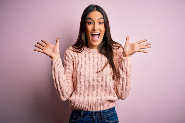 Young beautiful brunette woman wearing casual sweater over isolated pink background celebrating crazy and amazed for success with arms raised and open eyes screaming excited. Winner concept - Fotografie, Obrázek