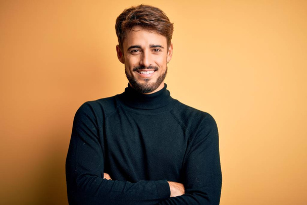 Fiatal jóképű férfi szakállal garbó pulóvert visel, sárga háttér felett áll boldog arc mosolyog keresztbe tett karral és a kamerába néz. Pozitív személy. - Fotó, kép