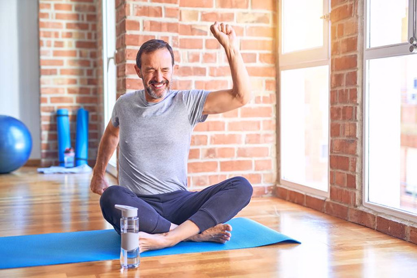 Middelbare leeftijd knappe sportman zittend op mat doen stretching yoga oefening in de sportschool Dansen gelukkig en vrolijk, glimlachend bewegend casual en zelfverzekerd luisteren naar muziek - Foto, afbeelding