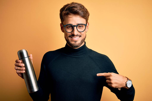 Young man on vacation wearing glasses holding metallic thermo wih water with surprise face pointing finger to himself - Foto, Imagem