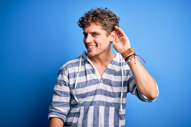 Young blond handsome man with curly hair wearing casual striped sweatshirt smiling with hand over ear listening an hearing to rumor or gossip. Deafness concept. - Photo, Image