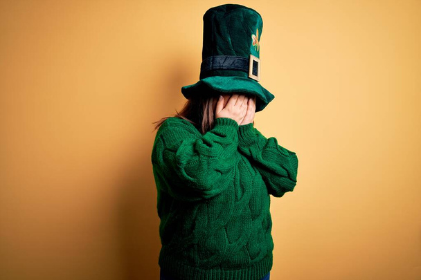 Молодая красивая женщина плюс размер носить зеленую шляпу с клевером празднует день святого Патрика с грустным выражением лица покрывая руки во время плача. Концепция депрессии
. - Фото, изображение