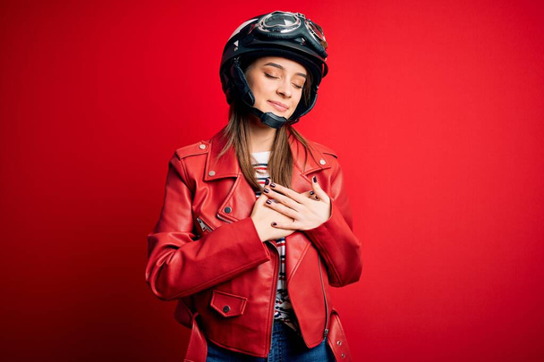 Young beautiful brunette motocyclist woman wearing motorcycle helmet and red jacket smiling with hands on chest with closed eyes and grateful gesture on face. Health concept. - Photo, Image