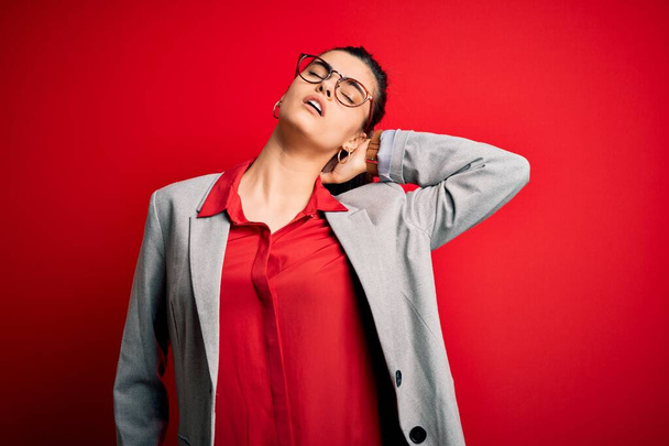 Молодая красивая брюнетка деловая женщина в пиджаке и очках на красном фоне страдает от травмы шеи, касаясь шеи рукой, мышечной боли
 - Фото, изображение