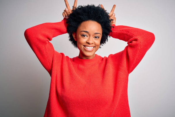 Jonge mooie Afro-Amerikaanse afro vrouw met krullend haar dragen rode casual trui Poseren grappig en gek met vingers op het hoofd als konijnenoren, glimlachen vrolijk - Foto, afbeelding