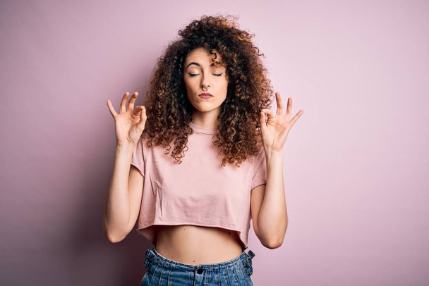 Young beautiful woman with curly hair and piercing wearing casual pink t-shirt relax and smiling with eyes closed doing meditation gesture with fingers. Yoga concept. - Foto, imagen