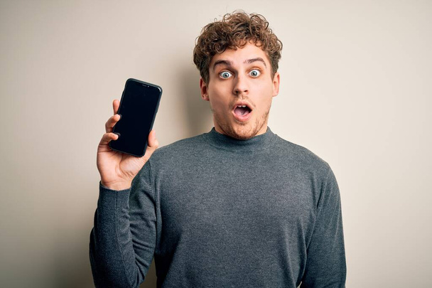 Mladý blonďatý muž s kudrnatými vlasy ukazující smartphone stojící nad izolovaným bílým pozadím vyděšený v šoku s překvapenou tváří, vyděšený a vzrušený výrazem strachu - Fotografie, Obrázek