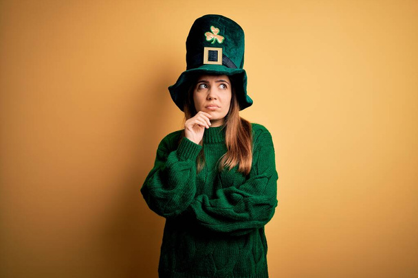 Young beautiful brunette woman wearing green hat on st patricks day celebration with hand on chin thinking about question, pensive expression. Smiling with thoughtful face. Doubt concept. - Photo, Image