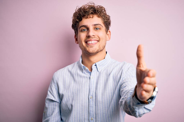 Young blond handsome man with curly hair wearing striped shirt over white background smiling friendly offering handshake as greeting and welcoming. Successful business. - Фото, изображение