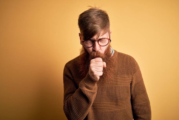 黄色の背景感の上に眼鏡と冬のセーターを身に着けているひげを持つハンサムなアイルランドの赤毛の男は具合が悪く、風邪や気管支炎の症状として咳をします。ヘルスケアの概念. - 写真・画像