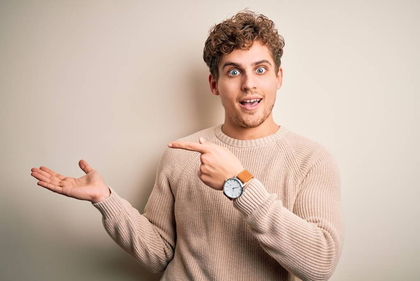 Young blond handsome man with curly hair wearing casual sweater over white background amazed and smiling to the camera while presenting with hand and pointing with finger. - Foto, Imagem