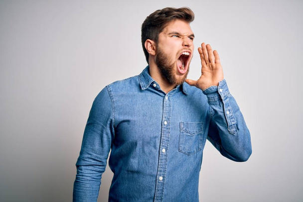 Young handsome blond man with beard and blue eyes wearing casual denim shirt shouting and screaming loud to side with hand on mouth. Communication concept. - Фото, изображение
