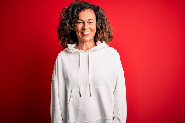 Middle age beautiful sporty woman wearing white sweatshirt over isolated red background with a happy and cool smile on face. Lucky person. - Photo, Image