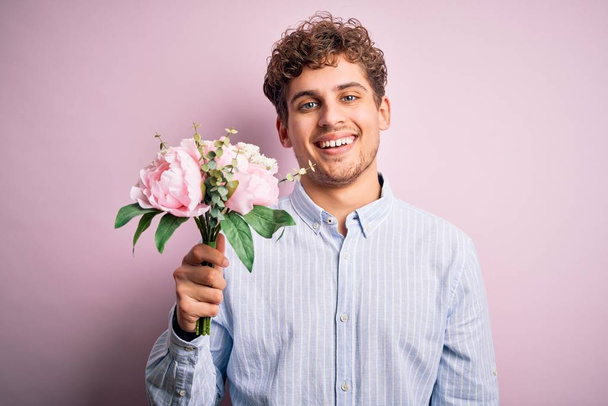 Young blond man with curly hair holding beautiful bouquet over isolated pink background with a happy face standing and smiling with a confident smile showing teeth - Фото, изображение