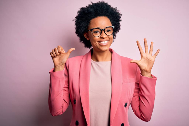 Junge schöne afroamerikanische Afro-Geschäftsfrau mit lockigem Haar trägt eine rosafarbene Jacke und zeigt mit Finger Nummer sechs nach oben, während sie selbstbewusst und glücklich lächelt. - Foto, Bild