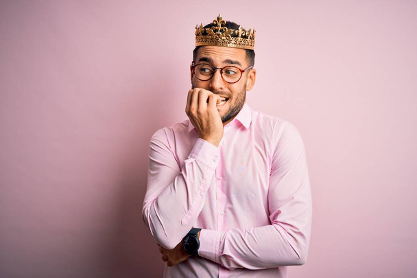 Fiatal jóképű üzletember visel arany koronát, mint király felett rózsaszín háttér úgy néz ki, stresszes és ideges a kezét a száj harapdáló körmök. Szorongás. - Fotó, kép
