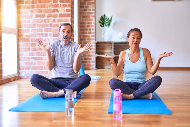 Sportliches Paar mittleren Alters, das auf einer Matte sitzt und Stretching-Yoga-Übungen im Fitnessstudio macht, ahnungslos und verwirrt mit erhobenen Armen und Händen. Zweifelhaftes Konzept. - Foto, Bild