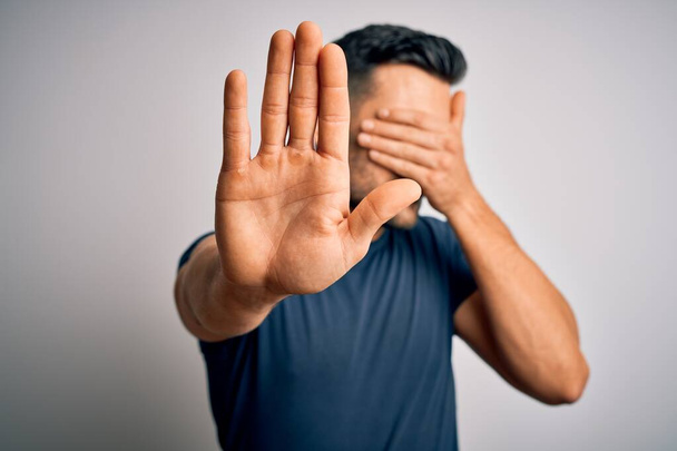 Jonge knappe man draagt casual t-shirt staan over geïsoleerde witte achtergrond bedekken ogen met handen en doen stop gebaar met droevige en angst uitdrukking. Beschaamd en negatief concept. - Foto, afbeelding