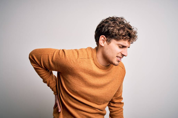 Νεαρός ξανθός όμορφος άντρας με σγουρά μαλλιά φορώντας casual πουλόβερ πάνω από λευκό φόντο Πόνος στην πλάτη, αγγίζοντας την πλάτη με το χέρι, μυϊκός πόνος - Φωτογραφία, εικόνα