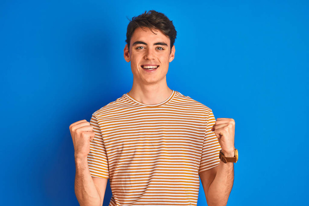 Teenager-Junge in lässigem T-Shirt steht vor blauem Hintergrund und feiert überrascht und erstaunt den Erfolg mit erhobenen Armen und offenen Augen. Siegerkonzept. - Foto, Bild