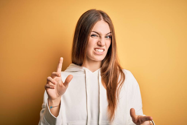 Young beautiful redhead sporty woman wearing sweatshirt over isolated yellow background disgusted expression, displeased and fearful doing disgust face because aversion reaction. With hands raised - Foto, imagen