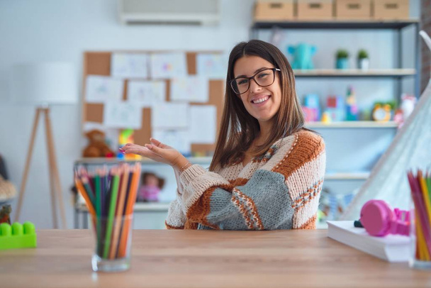 Молодая красивая учительница в свитере и очках сидит на столе в детском саду, указывая в сторону с открытыми руками ладонями, показывая копировальное пространство, представляя рекламу улыбаясь взволнованной счастливой
 - Фото, изображение