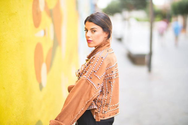 Νέο όμορφο κορίτσι φορώντας καφέ σακάκι στέκεται προς τα πίσω πάνω από πολύχρωμο τοίχο στο δρόμο της πόλης - Φωτογραφία, εικόνα