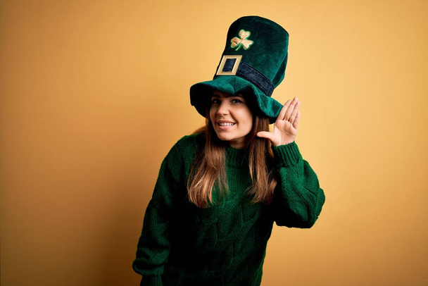 Jovem linda morena usando chapéu verde na festa do dia st patricks sorrindo com a mão sobre o ouvido ouvindo uma audição para boatos ou fofocas. Conceito de surdez
. - Foto, Imagem