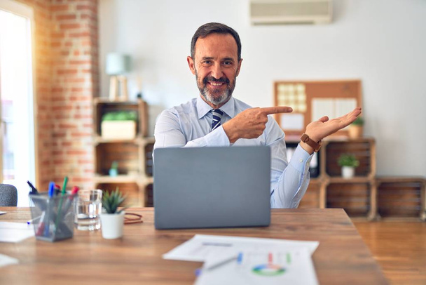 Μεσήλικας όμορφος επιχειρηματίας φορώντας γραβάτα κάθεται χρησιμοποιώντας φορητό υπολογιστή στο γραφείο έκπληκτος και χαμογελώντας στην κάμερα, ενώ παρουσιάζει με το χέρι και δείχνοντας με το δάχτυλο. - Φωτογραφία, εικόνα
