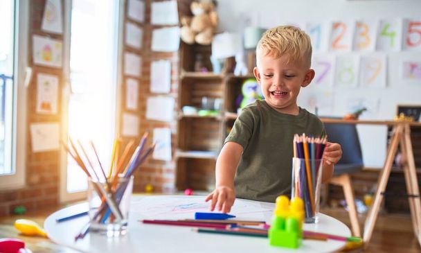 Νεαρό καυκάσιο παιδί παίζει στο νηπιαγωγείο ζωγραφίζοντας με χρωματιστά μολύβια. Παιδί του νηπιαγωγείου χαρούμενο στο playroom. - Φωτογραφία, εικόνα