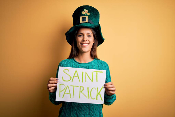 Zöld kalapos nő ünnepli a Szent Patrik napot, zászlót tart szent Patrick üzenettel, boldog arccal áll és mosolyog, magabiztos mosollyal a fogait mutatja. - Fotó, kép