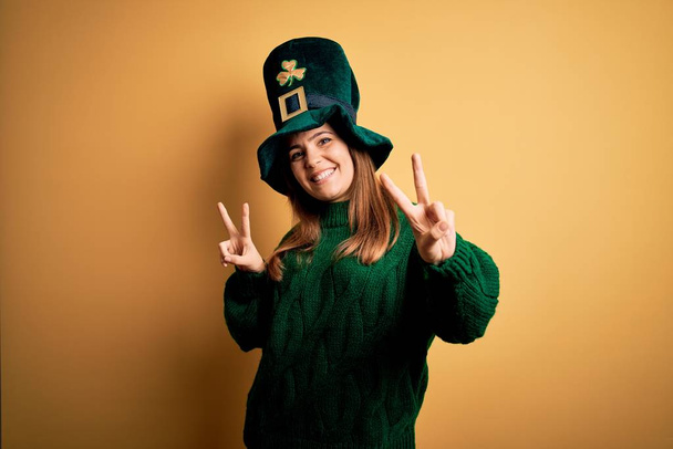Молодая красивая брюнетка в зеленой шляпе на праздновании дня святого Патрика улыбается, глядя в камеру, показывая пальцы, делающие знак победы. Номер два:
. - Фото, изображение