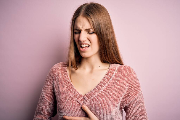 Young beautiful redhead woman wearing casual sweater over isolated pink background with hand on stomach because nausea, painful disease feeling unwell. Ache concept. - Photo, image