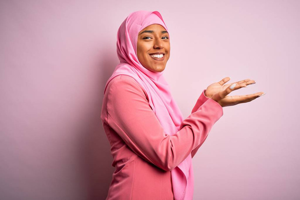 Młoda afro Afroamerykanka ubrana w hidżab muzułmański na odizolowanym różowym tle wskazując dłońmi otwarte dłonie pokazujące przestrzeń do kopiowania, prezentując reklamę uśmiechniętą podekscytowany szczęśliwy - Zdjęcie, obraz