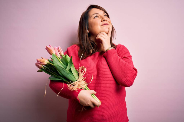 Belle femme plus taille tenant bouquet romantique de fleurs de tulipes naturelles sur fond rose avec la main sur le menton penser à la question, expression coûteuse. Visage souriant et réfléchi. Concept de doute
. - Photo, image