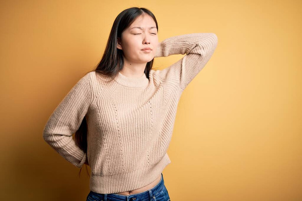 Young beautiful asian woman wearing casual sweater over yellow isolated background Suffering of neck ache injury, touching neck with hand, muscular pain - 写真・画像