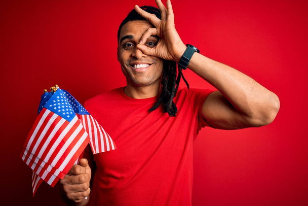 afrikanisch-amerikanischer Afro-Mann mit Dreadlocks trägt die Flagge der Vereinigten Staaten über rotem Hintergrund mit glücklichem Gesicht lächelnd tut ok Zeichen mit der Hand auf dem Auge, die durch Finger schaut - Foto, Bild